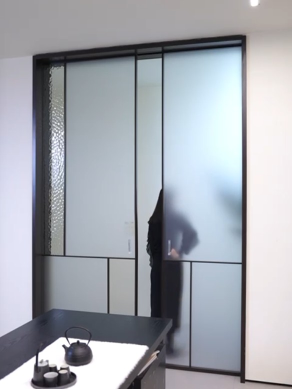 Black-framed sliding glass partition with an asymmetrical grid of frosted panels and a ripple-textured glass strip, a silhouetted figure moving behind, a tea set on a black table at the lower left
