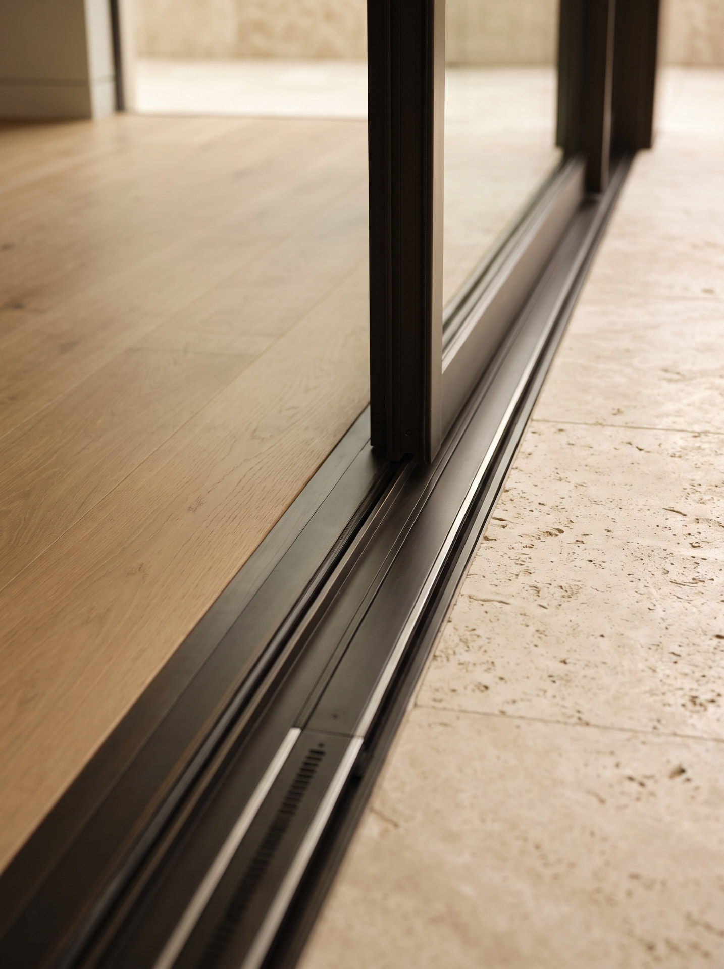 Low view of a dark bronze multi-track sliding door threshold set flush between light oak flooring inside and pale limestone pavers outside, with stainless runners and a drainage slot in the channel