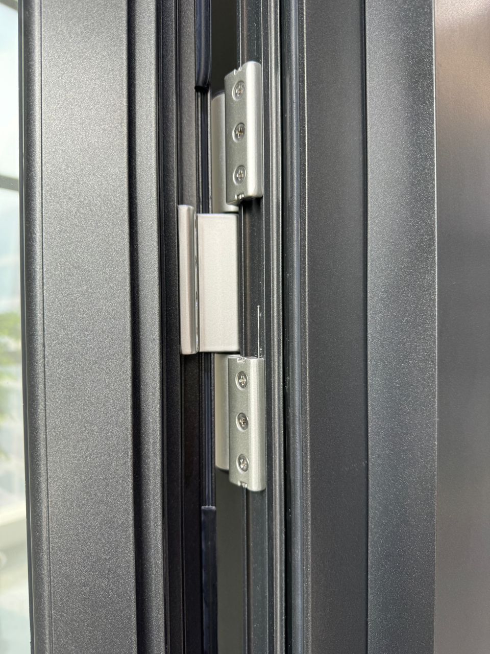 Extreme close-up of a satin metal hinge screwed into a textured charcoal frame channel beside a black rubber compression gasket