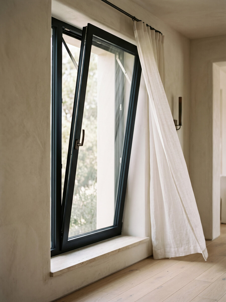 Window tilted open from the top in a warm plaster wall, sheer linen curtain beside a stone sill and pale oak floor