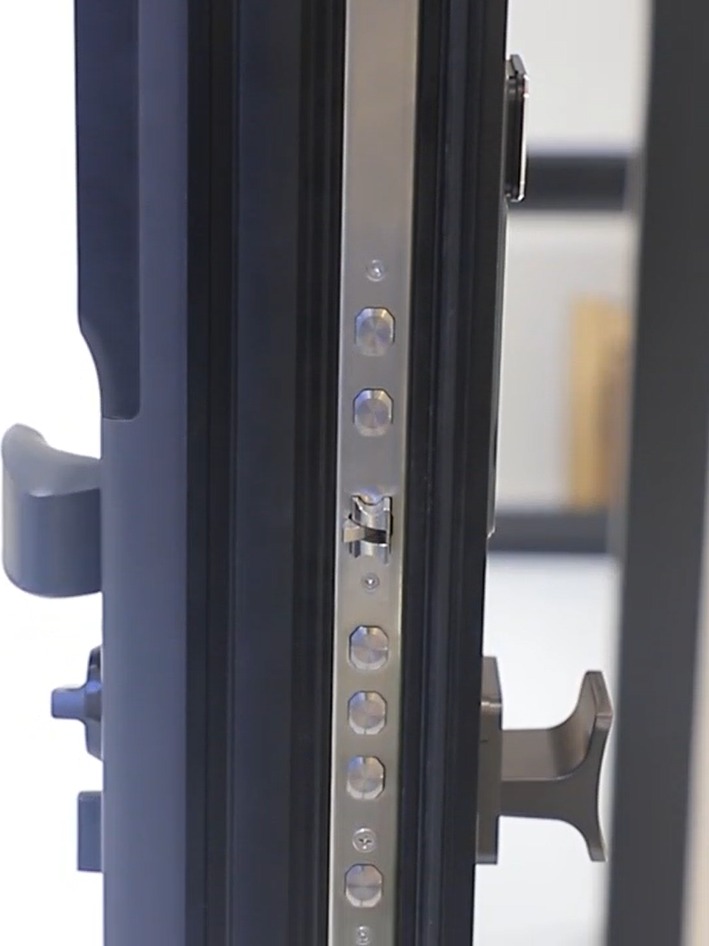 Close-up of the vertical edge of an open dark charcoal door frame showing a brushed stainless multi-point locking faceplate with several round bolts and a central latch, curved pull handles on both faces, a bright white background to the left and a softly blurred interior beyond