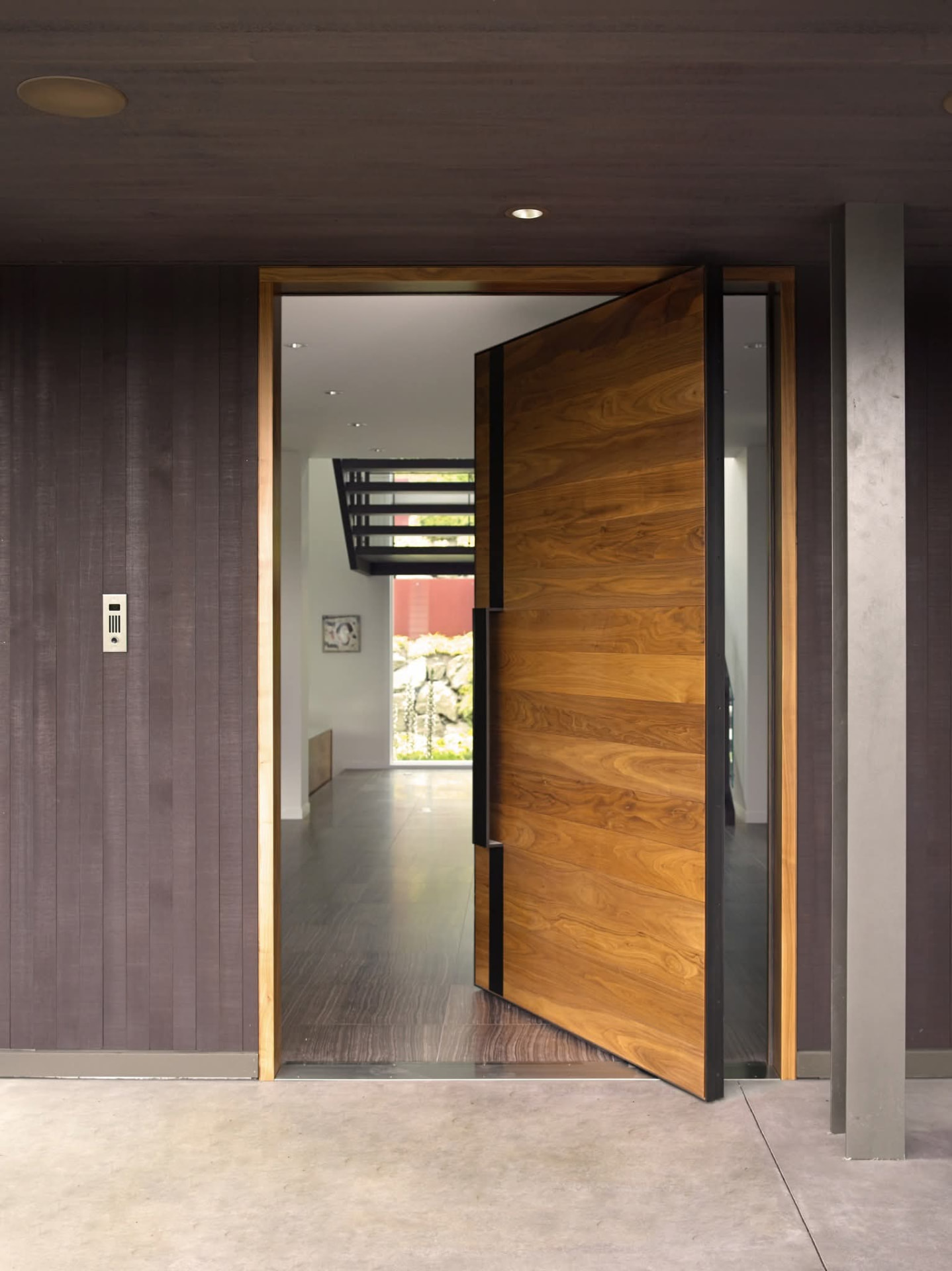 An oversized pivot entry door in horizontal honey-toned oak planks with a matte black steel frame and a full-height black pull handle, standing open into a bright foyer with an open-riser staircase beyond, dark vertical wood siding overhead and a raw steel column at the pale concrete porch