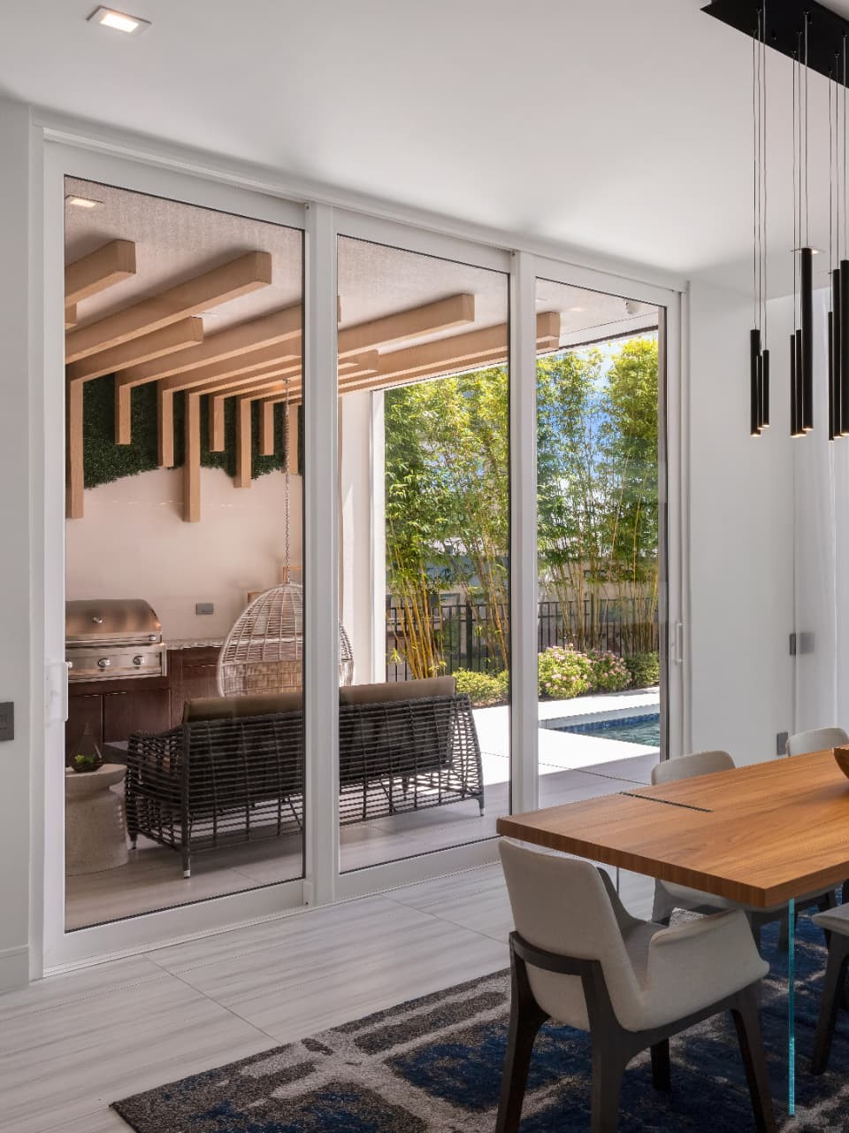 Open-plan dining room under black cylinder pendants with a whole wall of sliding doors opened to a covered patio, pool and lawn beyond