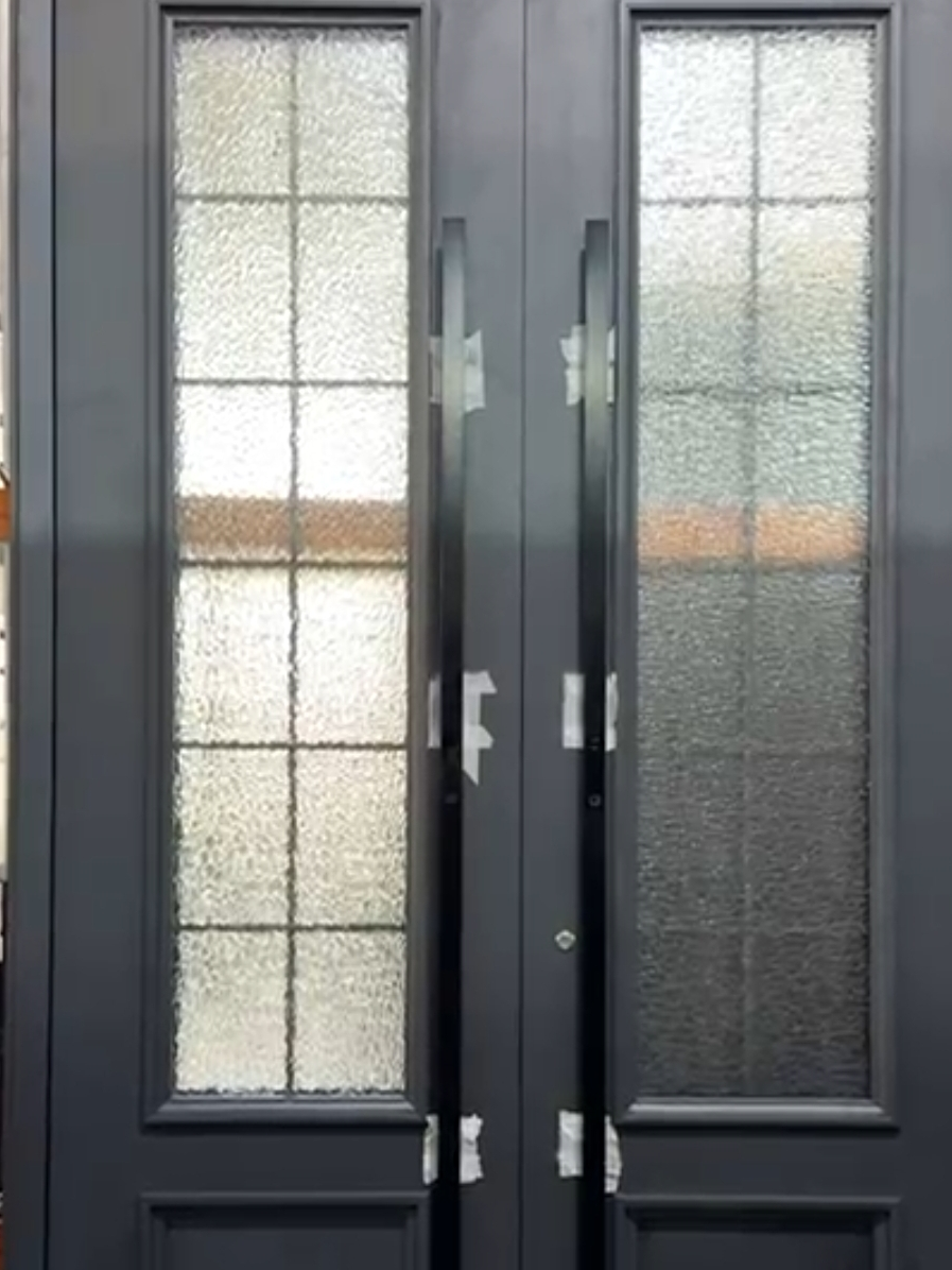 A pair of tall matte charcoal double entry doors with textured glass inserts divided by slender grid lines, long vertical black pull handles still taped from the shop, standing in a workshop before delivery