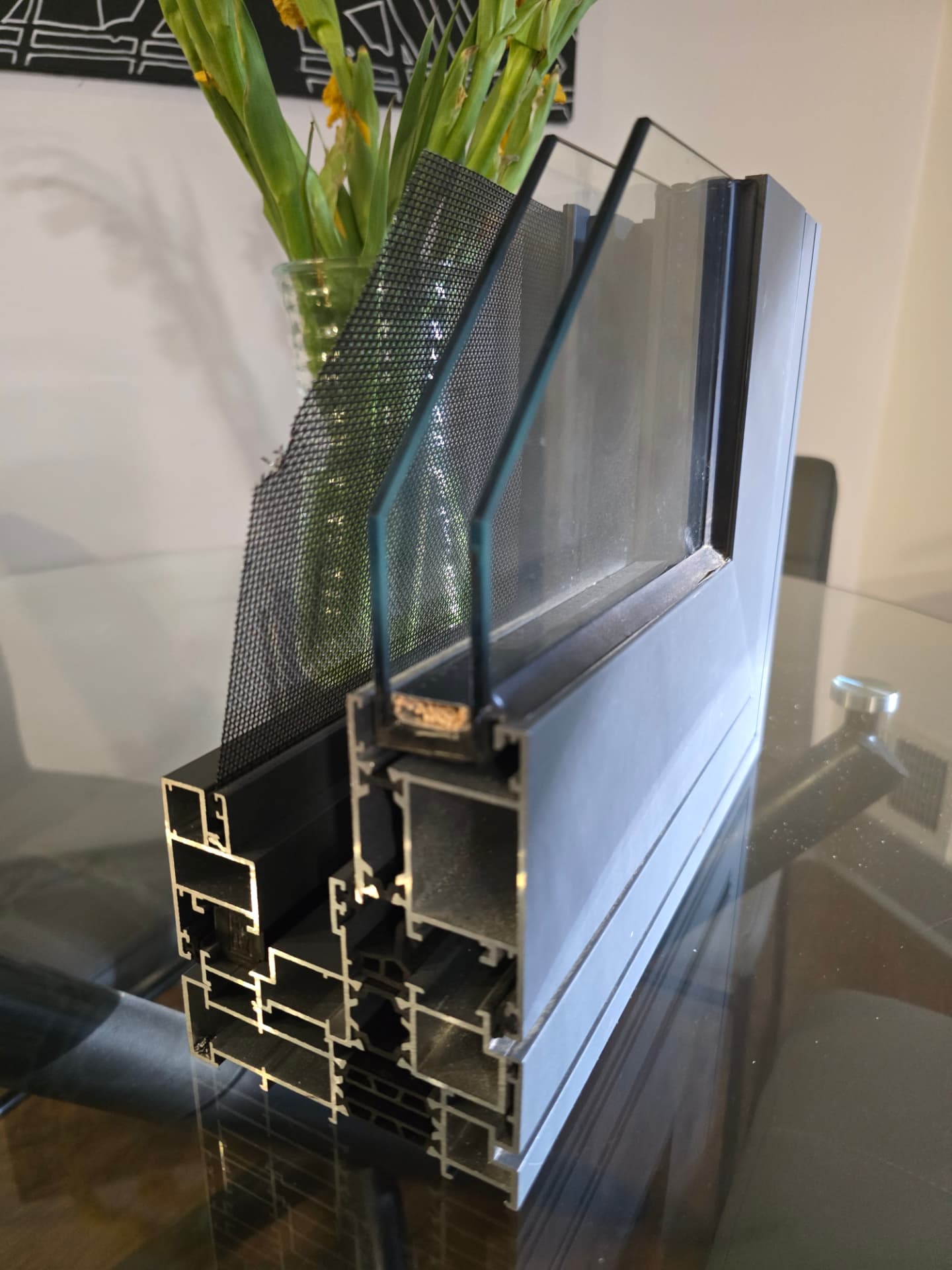 Cutaway sample of a charcoal window frame standing on a glass table, showing its chambered profile, double pane of glass and fine mesh screen, a vase of flowers soft in the background