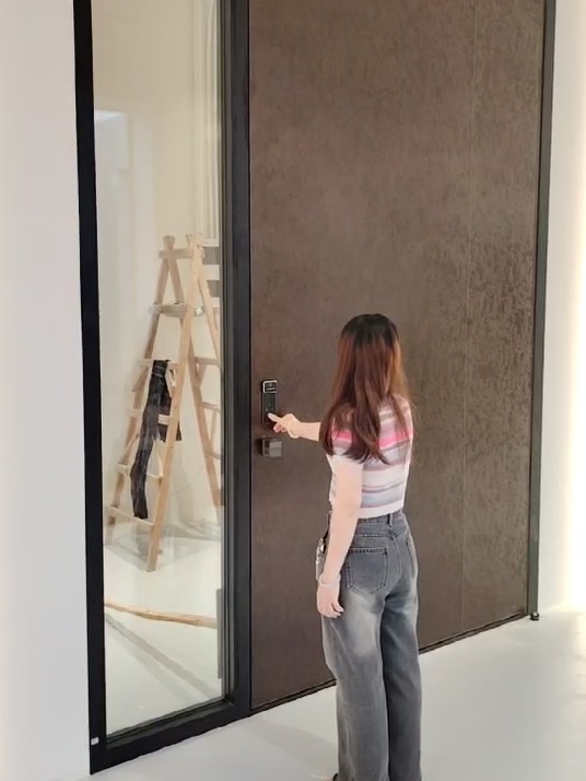 A woman seen from behind in a striped shirt reaching a tall matte black entrance door fitted with a vertical black smart-lock keypad and a square black pull handle, a full-height glass sidelight to the left, pale walls and a light tile floor