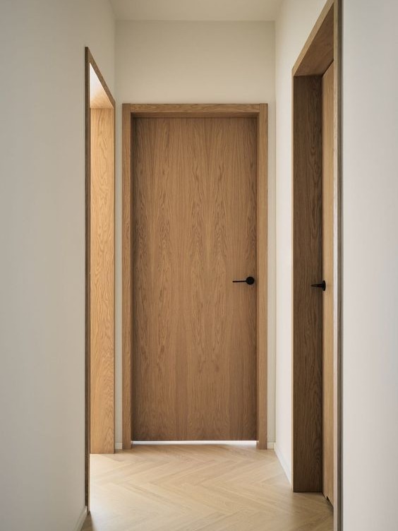 A modern interior hallway in warm oak with a closed flush door at the end, an open doorway with matching oak casing on the left, pale herringbone parquet flooring and slim matte black levers
