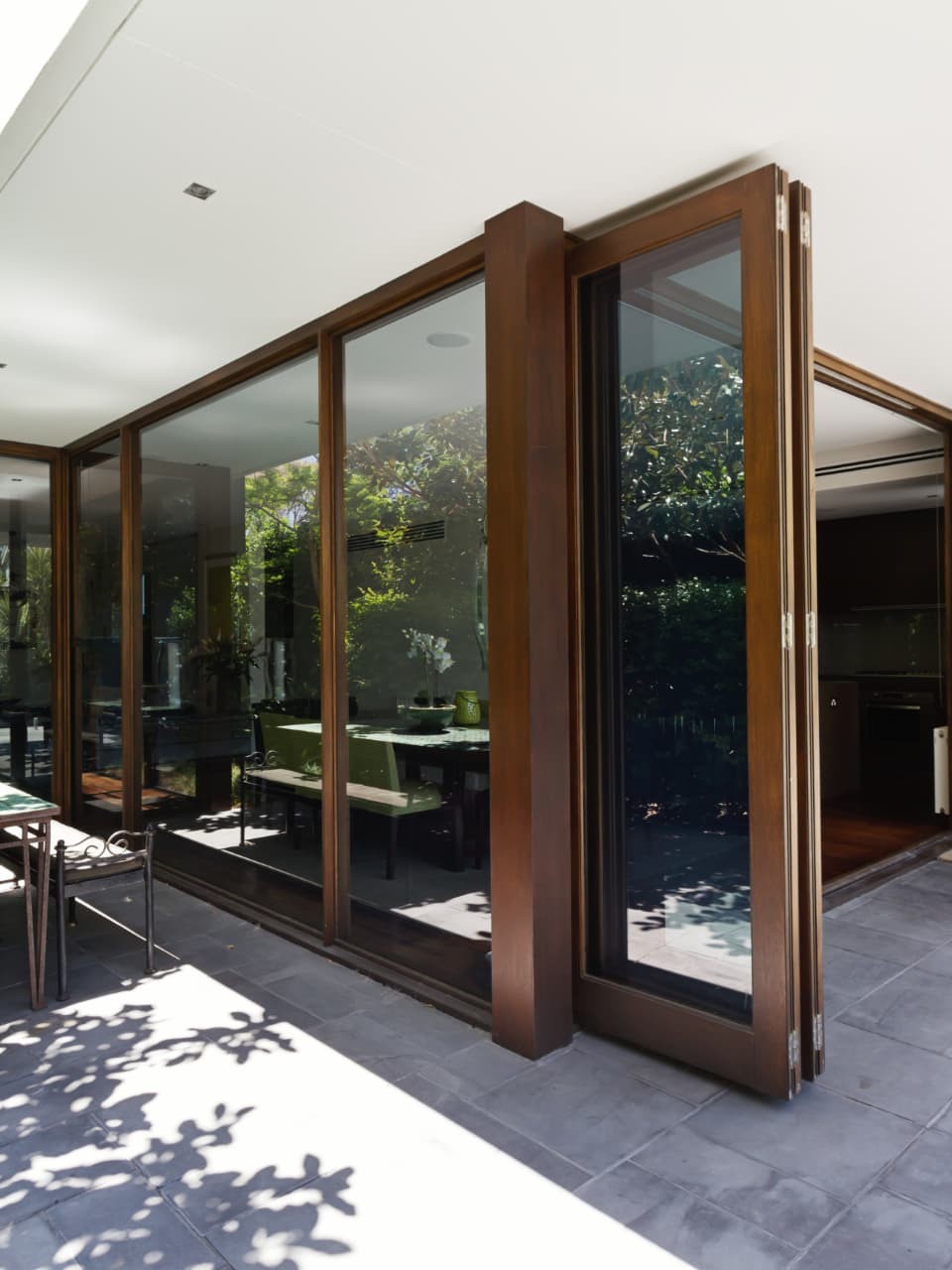Covered stone patio with dappled tree shadows and timber-framed folding glass doors opened at the corner to reveal a kitchen and dining room inside, an outdoor dining set at the left and a barbecue counter at the right