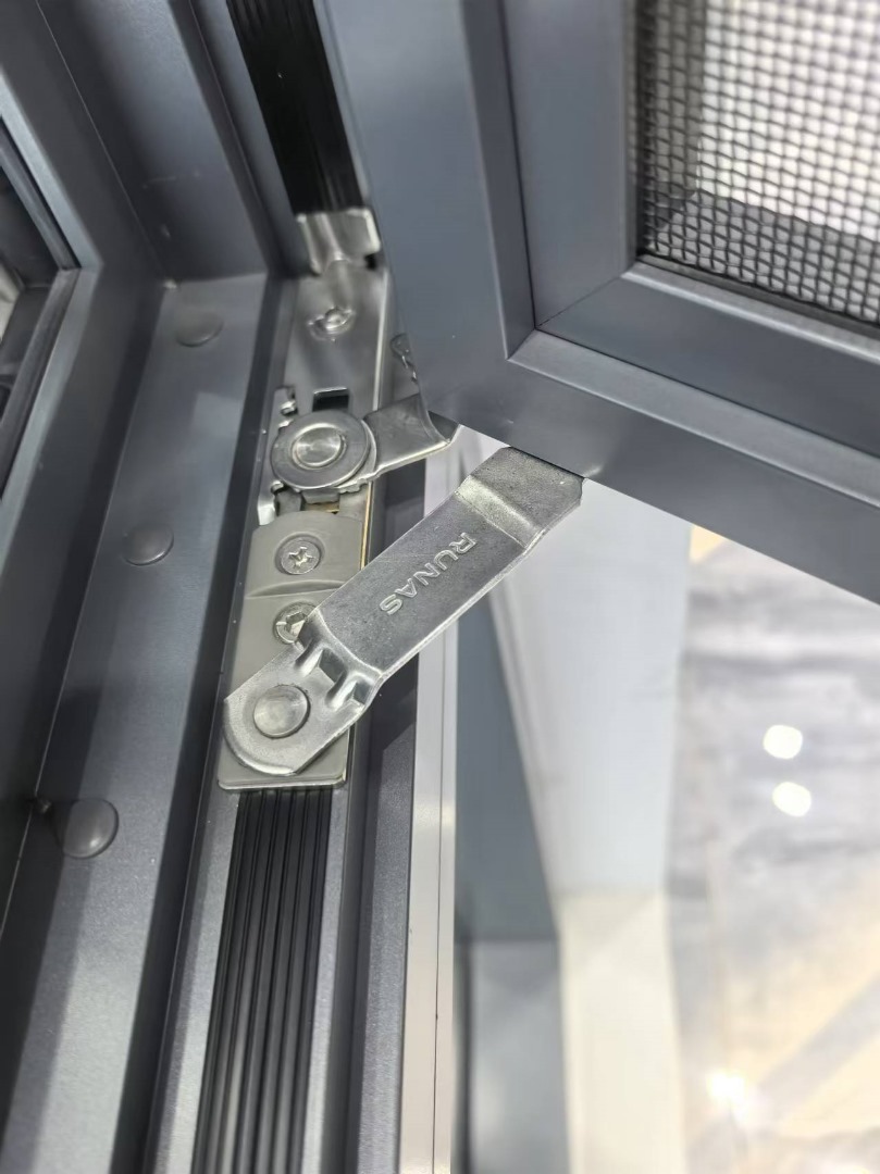 Open charcoal casement sash with a fine mesh screen and a brushed metal friction hinge arm holding it out, polished pale floor visible beyond the frame