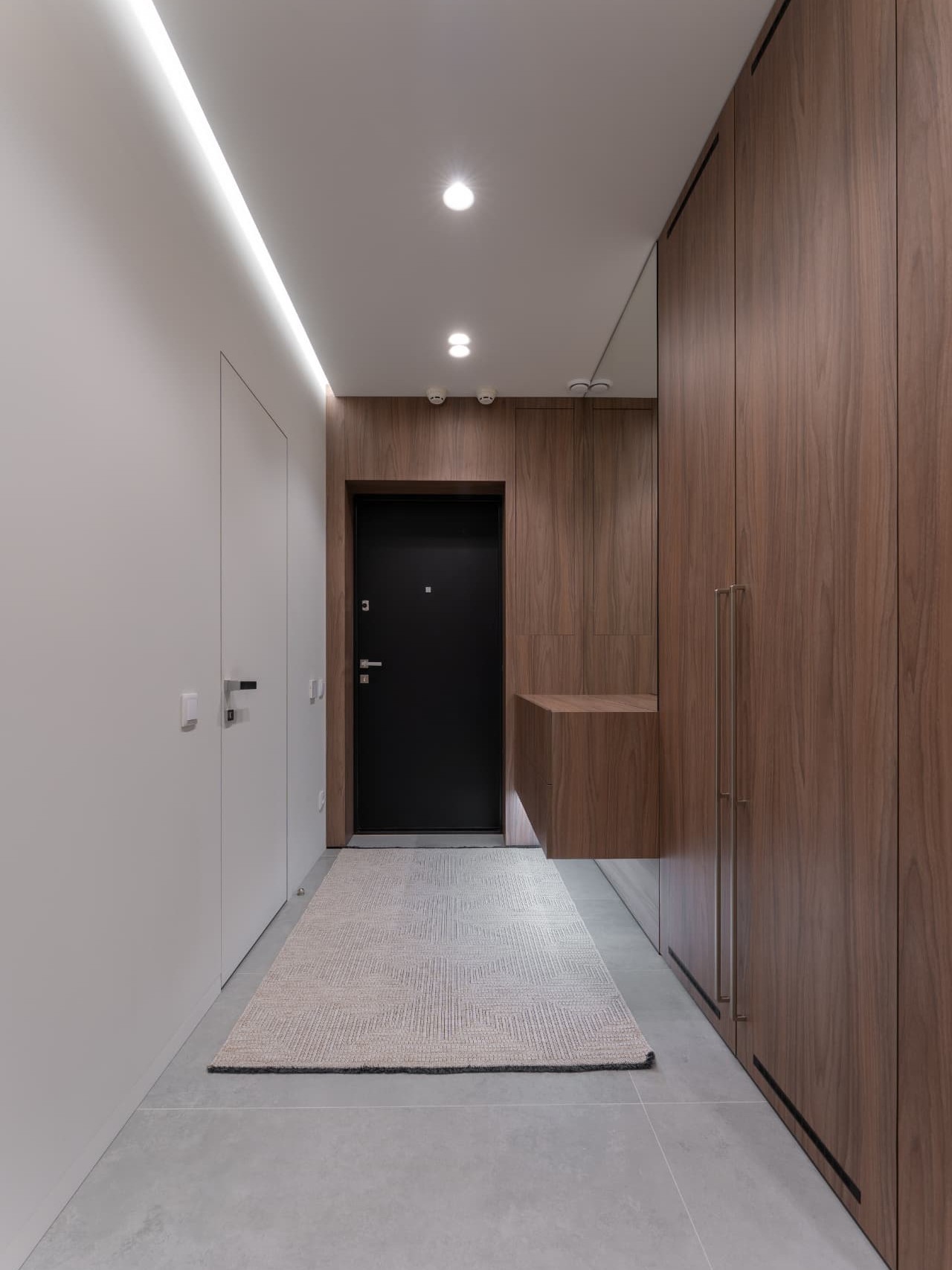 A bright residential entry hallway in one-point perspective: a seamless white wall with a flush interior door and slim silver lever on the left, floor-to-ceiling walnut-grain cabinetry with a mirror panel and a floating wooden console on the right, pale tiled floor with a woven runner leading to a matte black entrance door at the end