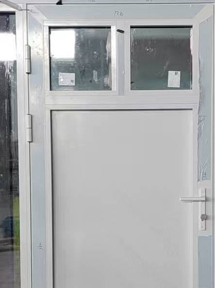 A white painted entry door slab with two small square glass lites at the top, two barrel hinges along the left jamb and a white lever handle on the right, still wrapped in protective film in the workshop