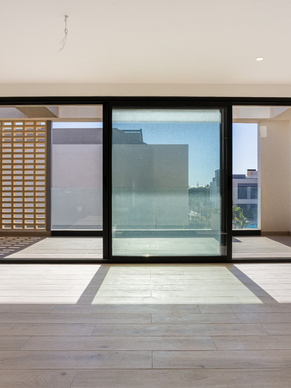 Unfurnished room in light wood and cream walls with floor-to-ceiling black-framed sliding doors opened onto a terrace behind a hollow-block screen